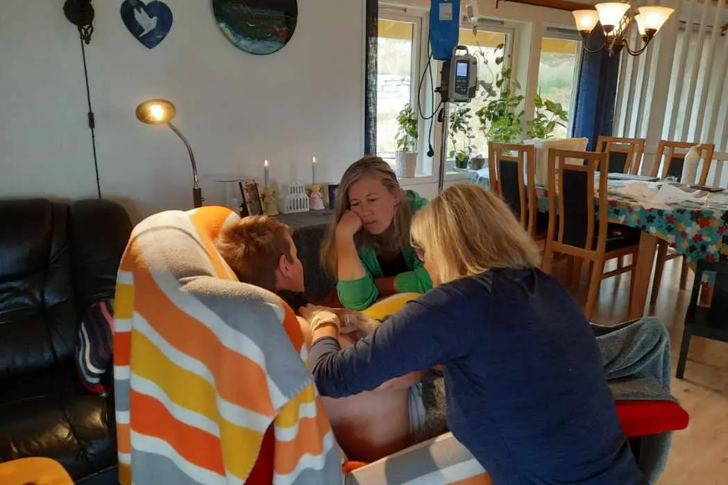 eit barn sit i ein stol og får behandling av ein sjukepleiar i heimen. Ei kvinne med grøn gensar sit ved sidan av barnet.