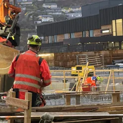 Ein mann iført arbeidskle jobbar på ein byggeplass. I bakgrunnen ein grabb frå ei gravemaskin og delar av eit bygg