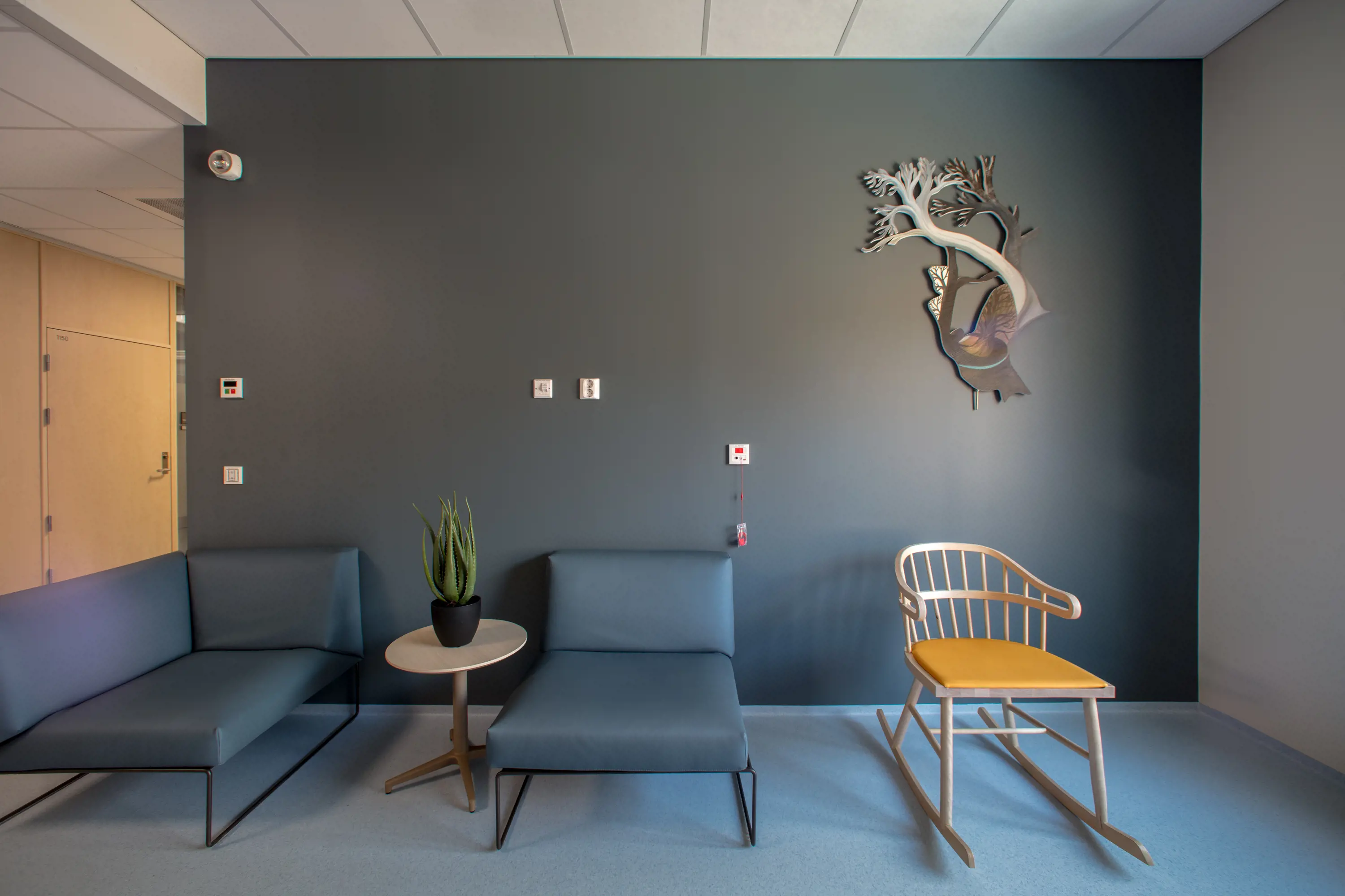 eit miljø med brun vegg med eit kunstverk i tre på veggen, og to stolar i blått, samt ein trestol 
