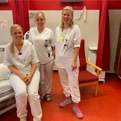 tre kvinnelege sjukepleiarar som er kledd i kvit uniform inne på eit pasientrom med raude gardiner og møblement.