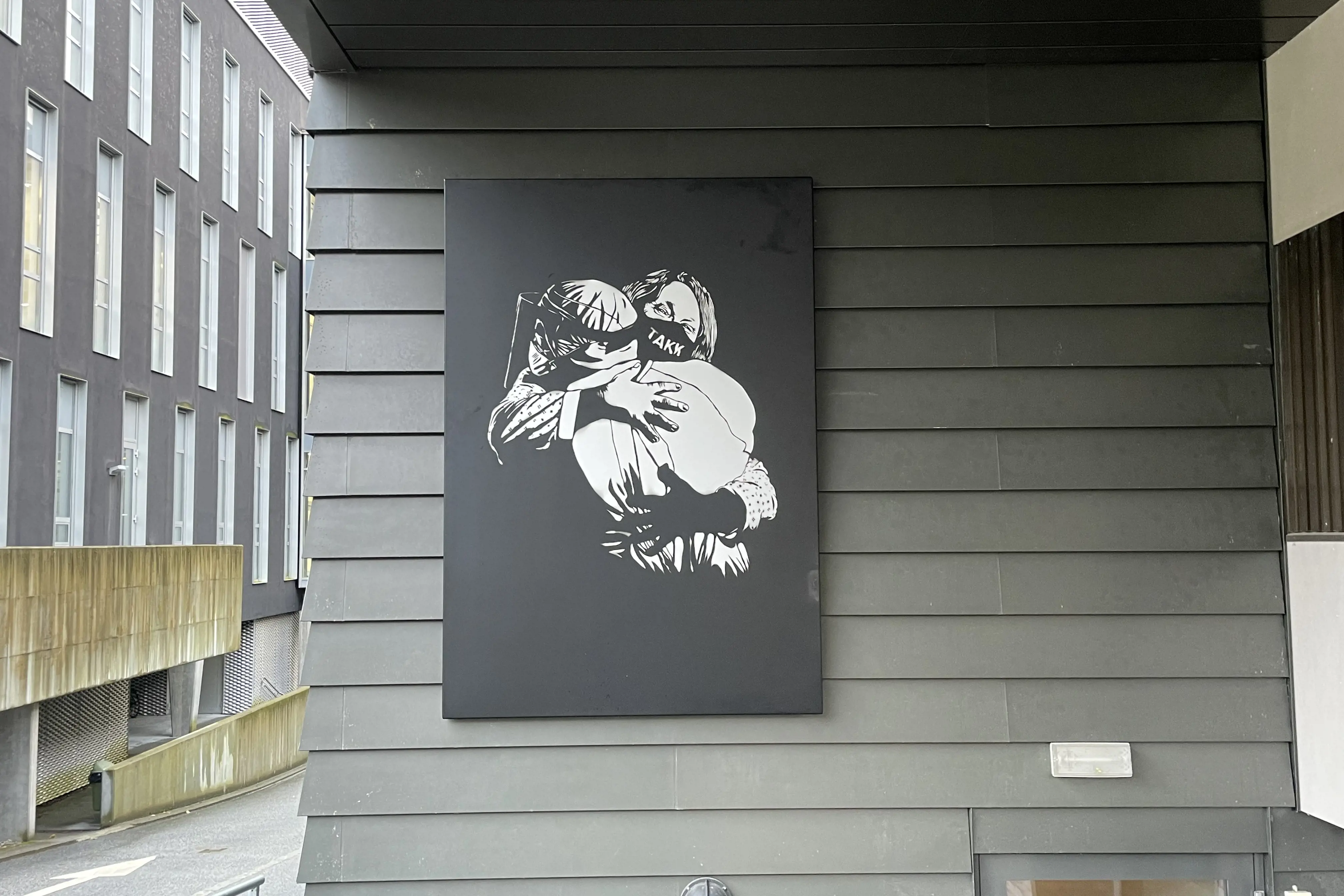 kunstverk som heng på ein grå vegg med panel utandørs av to personar som omfavnar kvarandre